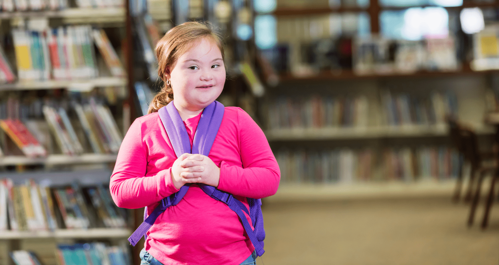 petite fille adorable en pull rose qui tient les anses de son cartable, elle est debout dans une bibliothèque scolaire