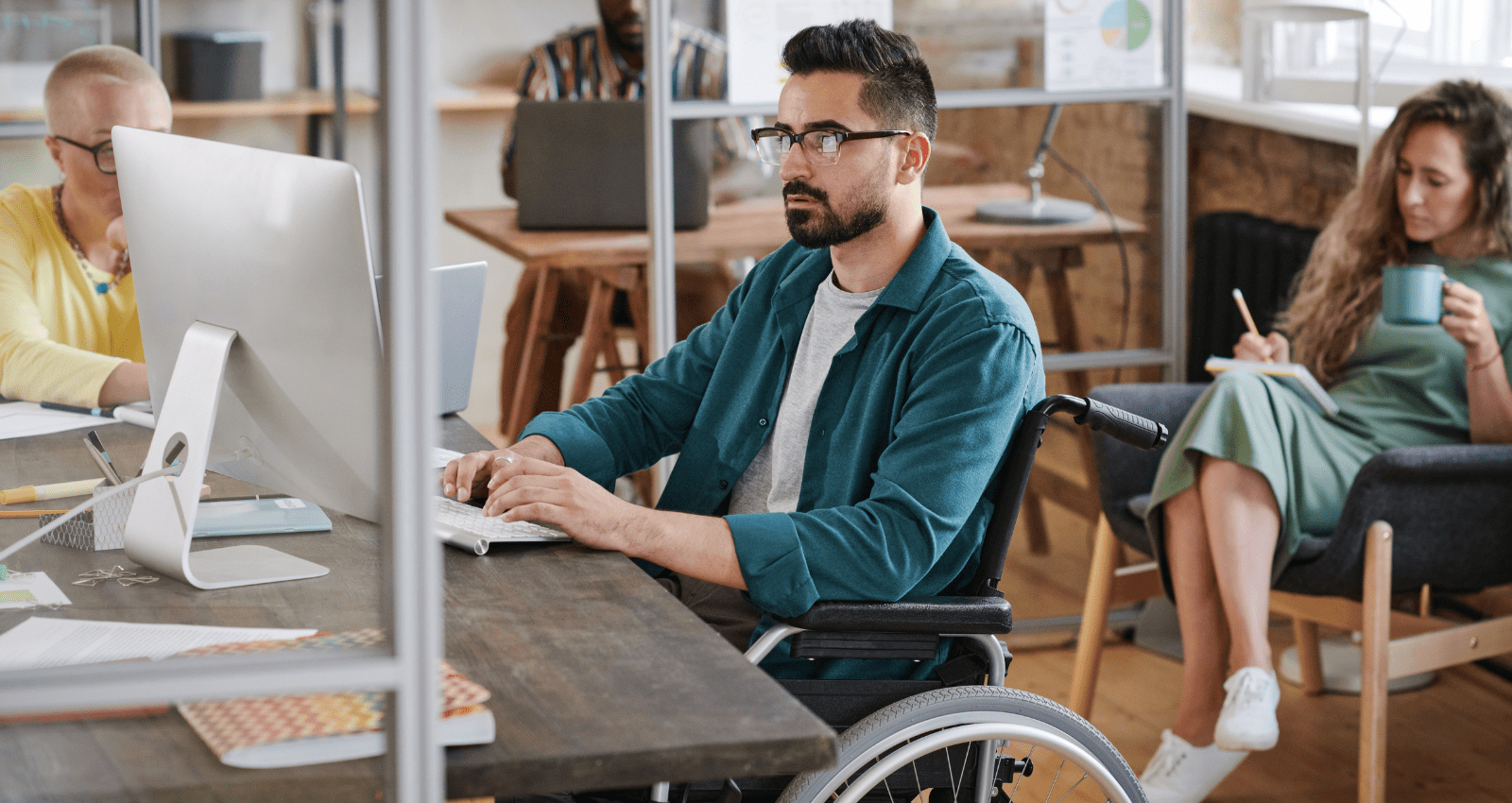 homme en fauteuil rouant assis à son bureau dans un open space avec d'autres collègues